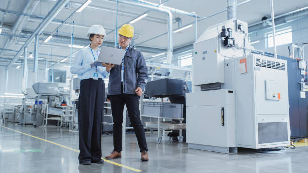 istock Two Young Asian Heavy Industry Engineers in Hard Hats Walking with Laptop Computer and Talking in a Factory. Two Manufacturing Employees at Work in Research and Development Facility. Medium Angle Shot 1462149163