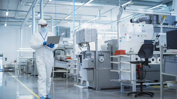 重工業製造工場:ラップトップコンピュータで歩く滅菌カバ��ーオールの科学者、産業用cncマシンの設定を調べ、生産機能を構成する。 - 製薬業 ストックフォトと画像