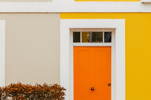 The colorful pastel yellow facade with orange front door of a traditional terraced London mews townhouse.
