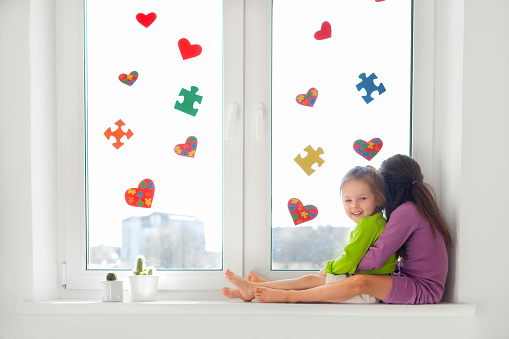 Two girls are sitting on the windowsill by the window decorated with hearts with colored puzzles inside - a symbol of autism. Support and care for loved ones