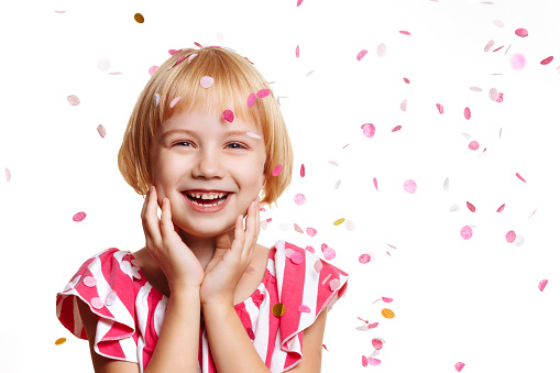 A girl in a festive dress on a spruce background laughs holding her hands to her face, confetti fly around. Concept for birthday party, greeting cards and holiday social media posts.