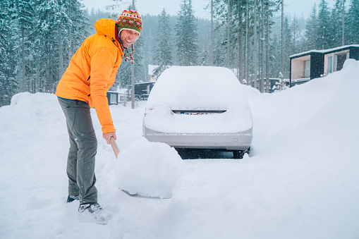 Winter morning routine. Smiling man with a shovel removing snow from the path and cleaning auto. Car covered with snow as a huge snowdrift on the countryside forest home driveway.