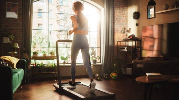 Beautiful Athletic Sports Woman Running on a Treadmill at Her Home Gym. Energetic Workout: Female Training while Listening Podcast or Music in Headphones. Apartment with Window View. Side Back View hermosa mujer deportiva atlética corriendo en una cinta de correr en el gimnasio de su casa. entrenamiento energético: entrenamiento femenino mientras escucha podcast o música con auriculares. apartamento con vistas a la ventana. vista lateral posterior