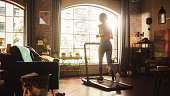 Beautiful Athletic Sports Woman Running on a Treadmill at Her Home Gym. Workout Female Athlete Training while Listening Podcast or Music in Headphones. Apartment with Window View. Side Back View