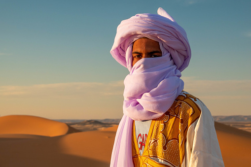 A young Berber man wearing a large lilac turban in the Chebbi desert in Morocco. He looks into the lens of the camera. In the background the orange coloured dunes of the Chebbi desert