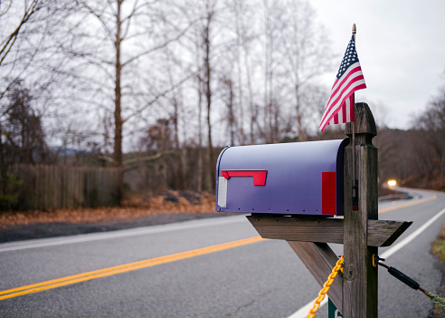A roadside lilac mailbox and protruding Stars and Stripes flag by a tarmac road with winter trees and an oncoming vehicle in the far distance.