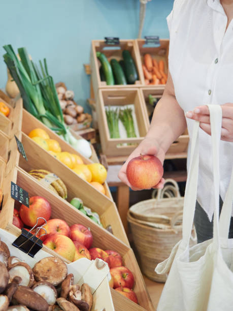 Partial view of an unrecognizable customer choosing an apple in a grocery store with a reusable cloth bag. Healthy and sustainable shopping concept in a small local store.
