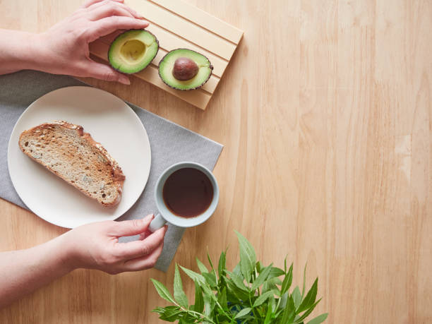 Woman's hands holding coffee cup and picking up half avocado on breakfast table. Top view with copy space.