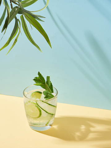 Summer drink in crystal glass with lime, cucumber and mint on yellow and blue background with leaves and hard shades. Cold mocktail non-alcoholic soda water and mint tonic. Summer color concept.