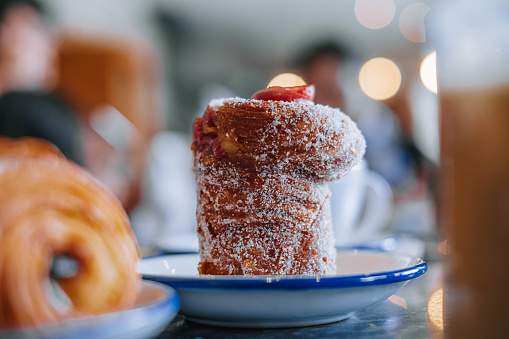 A close-up shot of a delectable Cruffin or Kraffin pastry in a small white plate. The focus is on the pastry, showcasing its golden-brown, flaky crust and soft, fluffy center. The background is blurred with a bokeh effect, emphasizing the pastry and creating a mouth-watering visual for the viewer.