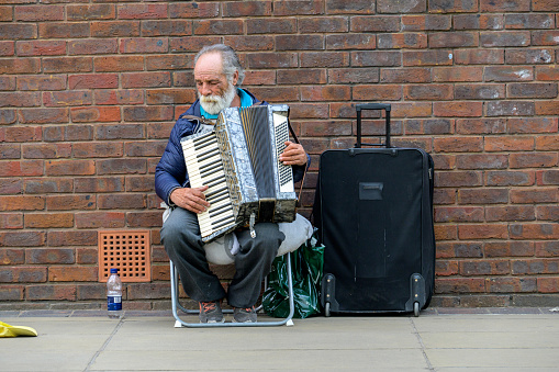 LONDON - May 21, 2022: Elderly busker with white beard plays accordion next to old red brick wall