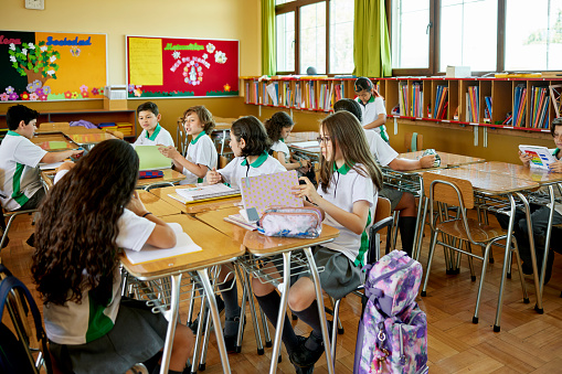 Elementary-age boys and girls in uniforms seated, having just arrived, opening their notebooks, and conversing.