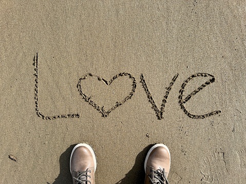 love, hand written love word on sand. female feet or woman standing on a sandy beach next to heart. romance on beach with copy space. romantic relationship or valentines day concept