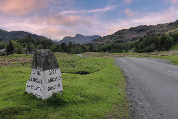 Milestone outside the Lake District village of Elterwater with the Langdale Pikes in the distance English Lake District, Cumbria, England, UK - Old fashioned milestone alongside road outside Elterwater village with the mountain peaks of the Langdale Pikes in the distance viewed against a sunset sky langdale pikes stock pictures, royalty-free photos & images
