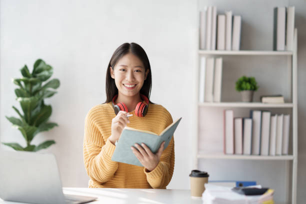 istock Young adorable Asian woman writhng on notebook while doing on her homework for the class. Doing homework at home comfortable. 1462213007