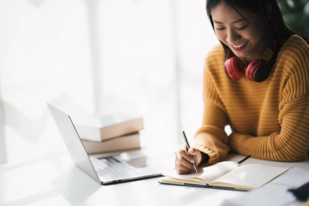 istock Young adorable Asian woman writhng on notebook while doing on her homework for the class. Doing homework at home comfortable. 1462214372