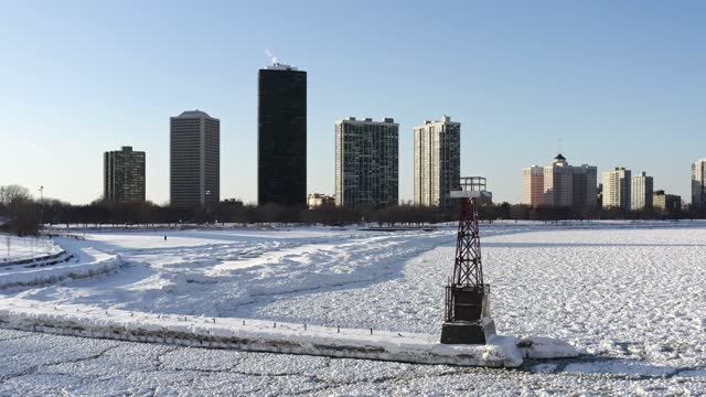 Panning out aerial from a snow and ice covered pier and Lake Michigan shoreline in Chicago as a person walks across the beach with high-rise residential buildings in the Edgewater neighborhood beyond.