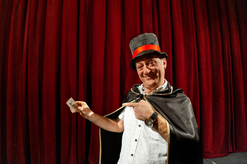 Portrait of the senior male magician, wearing a tail coat and cylinder while performing magic tricks on the stage at the theatre