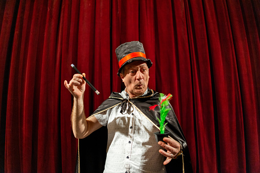Portrait of the senior male magician, wearing a tail coat and cylinder while performing magic tricks on the stage at the theatre