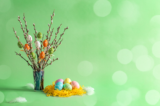 Easter eggs decorations, feathers and pussy willow twigs with catkins in a vase. Beautiful defocused lights bokeh. Space for copy.