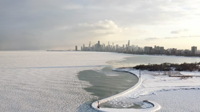 Panning out and lowering down aerial drone footage of the Chicago skyline with an ice and snow covered curved pier and Lake Michigan coming into view in the foreground.