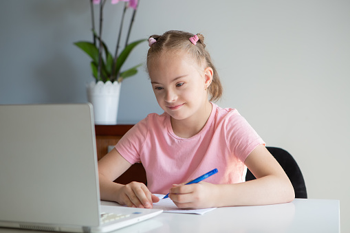 A girl with Down syndrome does her homework at home, prepares her hand for writing, prepares to go back to school. Working out while sitting at a laptop