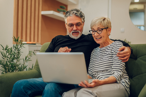 Portrait of a senior married happy couple embracing using laptop together and sitting on a couch at home. Smiling elderly spouses reading news, shopping online. Older people and computer concept.