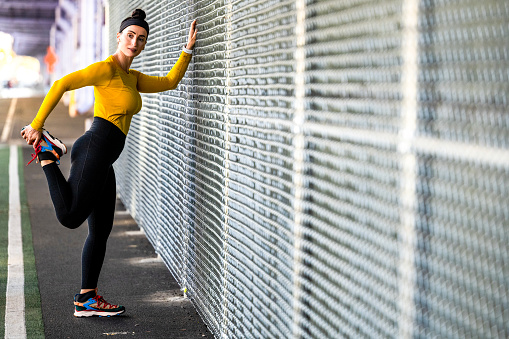 A fit woman in yellow t-shirt, black leggings and colorful sport shoes with wireless headphones and a smart watch seen stretching by the fence after a great running session during her weekend fitness activity in new York City.