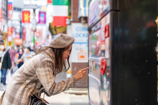 Happy Asian woman choosing and buying drinks on snack and beverage vending machine while shopping at shibuya, Tokyo, Japan. Attractive girl enjoy and fun outdoor travel city street on autumn vacation.