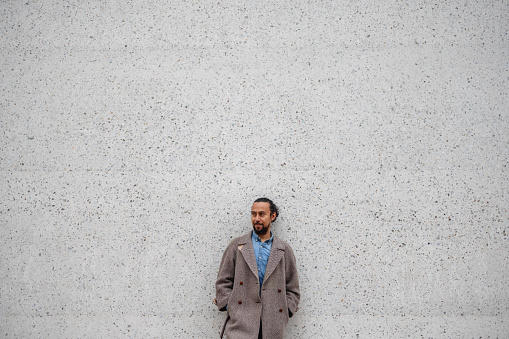 Portrait of latin adult male with dark hair and dark beard leaning back on large grey wall and looking sideways. He is wearing casual outfit.