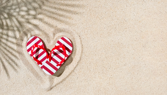 Pare of red flip-flops in shape of heart on sand with palm leaves shadow, copy space, tropical Valentine concept