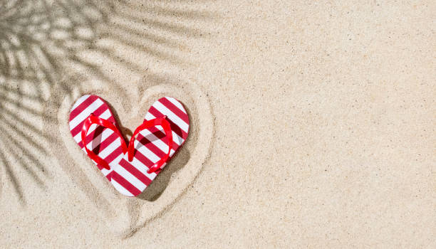 Pare of red flip-flops in shape of heart on sand with palm leaves shadow, copy space, tropical Valentine concept stock photo