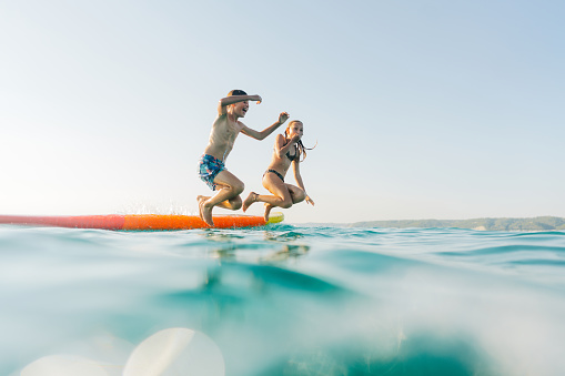 Photo of two kids having fun while spending summer by the sea, jumping of the paddle board