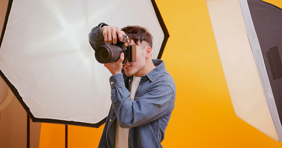 asian young male photographer holding professional camera for shooting standing in front of yellow background at studio