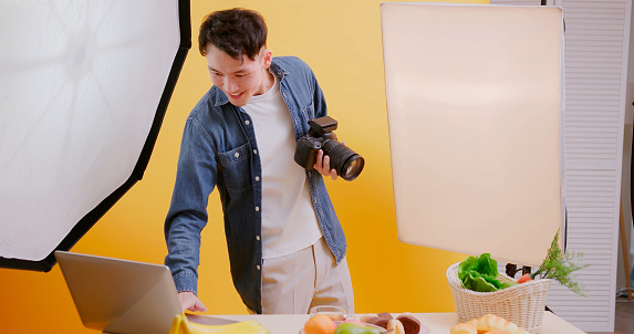 asian young male photographer use professional camera shooting food in front of yellow background at studio