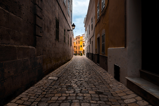 Carrer d'es Mirador in Ciutadella de Menorca showcases idyllic yellow townhouses, cobbled lane and decorative facades along a narrow traditional alleyway, illuminated with daylight against cloudy sky.