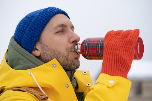 Attractive satisfied English man in winter warm clothes drinks hot tea from checkered thermos breathe air. Peaceful Scandinavian man holding thermo bottle in hand near lips outdoors in cold weather.