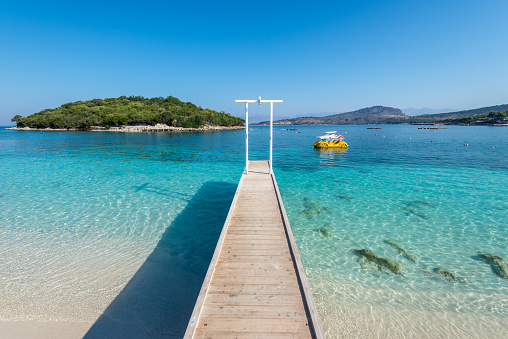 Beautiful sandy Ksamil beach with wooden walkway with shower in Albania. Idyllic little island surrounded by incredible turquoise lonian sea in the background.