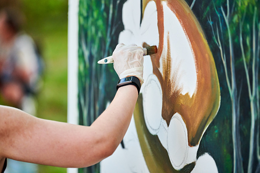 Girl artist hand holds paint brush and draws green nature landscape on white canvas at outdoor art painting festival, paintings art picture process. Woman artist paints atmospheric surreal picture
