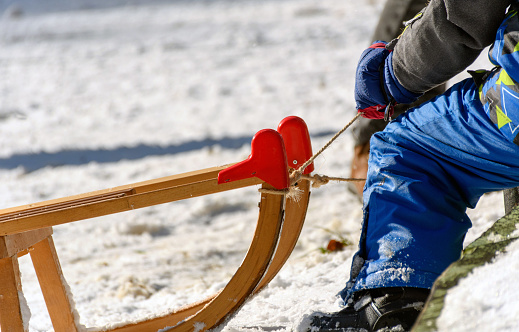 Kid wearing outdoor winter clothes, sitting on wooden sled, ready to ride downhill on snowy slope on a sunny winter day