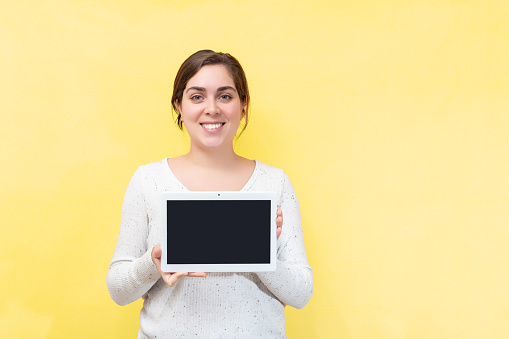 A Caucasian Smiling positive young woman shows a blank screen tablet isolated with a yellow wall