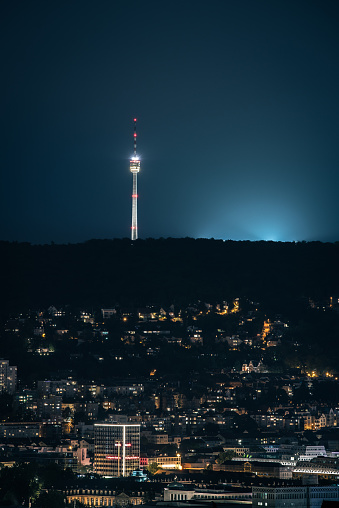 A vertical shot of an illuminated city and a telco tower against a blue nigh sky