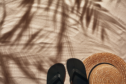 Summer concept with a shadow of a tropical palm tree leaves, copyspace. Traveler vacation accessories such as black thongs and a straw hat are laid out on a perfect beach sand. Summertime lifestyle, objects in flat lay, top view arrangement.