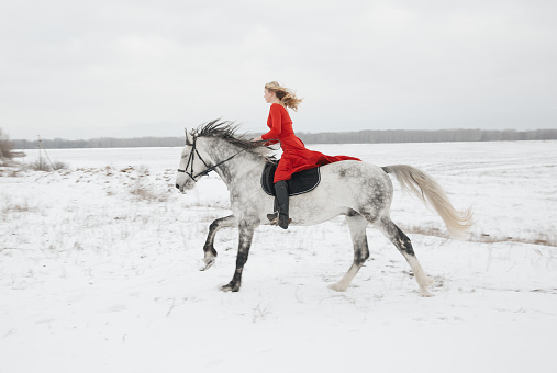 Woman jockey in a red dress rides a fast galloping horse - snow winter fields view