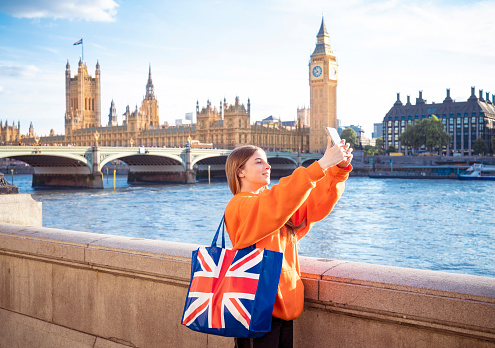 London blond tourist young student woman with UK flag shopping bag  shooting selfie at Big Ben and Houses of Parliament in Westminster bridge at sunset with red bus in UK Great Britain United Kingdom, England at the evening dusk