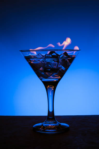 brennendes alkoholisches getränk mit eiswürfeln, auf blauem farbverlaufshintergrund, vertikaler rahmen. brennender cocktail auf dem tisch in einer bar - martini brand vermouth stock-fotos und bilder