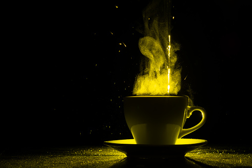 A mug with a warm drink and steam illuminated by yellow light, copy space, creative. Steaming coffee cup on a black background, silhouette. Morning coffee