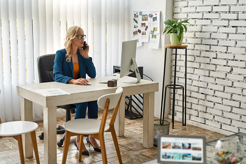 Businesswoman talking on smartphone and watching computer at desk in spacious office. Concept of modern successful female lifestyle. Young concentrated blonde caucasian woman wearing suit and glasses