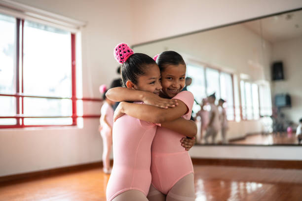 Two young female ballerinas, fair-haired and white-skinned, elegantly rehearse ballet poses at barre. Studio setting features soft lighting and light wooden flooring, enhancing serene atmosphere