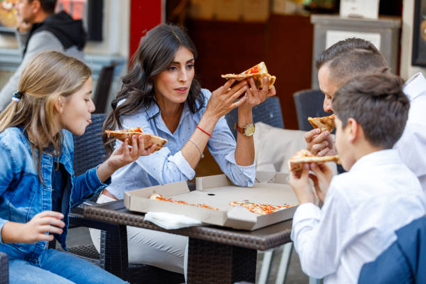 irmãozinho e irmã estão felizes em comer uma fatia de pizza com seus pais na cidade. - little girls group of people happiness cheerful - fotografias e filmes do acervo